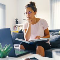 Woman looking at online course on her laptop and taking notes in a book on her lap.