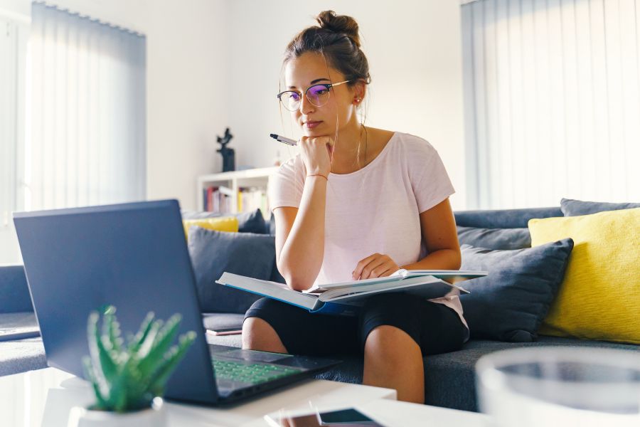 Woman looking at online course on her laptop and taking notes in a book on her lap.