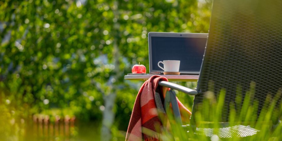 Chair placed outside surrounded by greenery, with laptop, coffee cup, and apple nearby