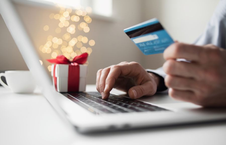 A person's hands holding a credit card and typing the information into a nearby latptop. A neatly wrapped gift is next to the computer.