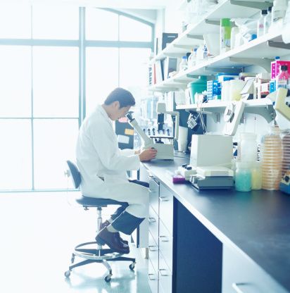 Researcher in a white lab coat sitting at a lab bench looking into a microscope.