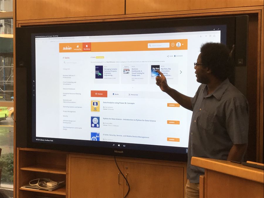 A training specialist in front of a large monitor in the Library Commons, helping people learn a new application during Tech Tuesday.