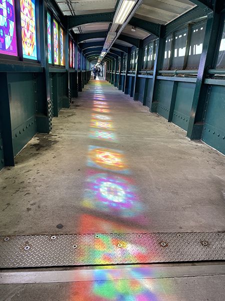 Light streams through the stained glass windows of a subway platform, leaving colorful patterns on the concrete floor.