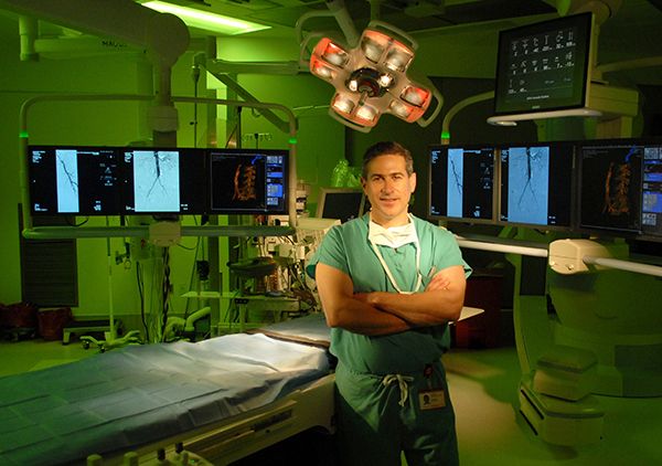 A surgeon wearing hospital scrubs standing in an operating room with a green light behind him.
