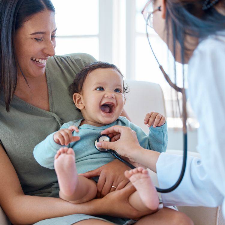 A baby being held by their mother while a pediatrician listens to their heartbeat.